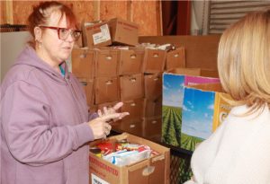 Two people in a warehouse with boxes of food. One gestures while speaking. The setting appears to be a food distribution area.