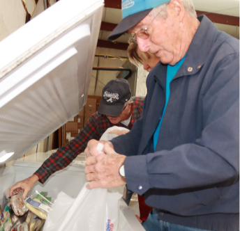 Two people are organizing frozen food items into bags from a large open freezer in a storage space.