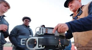 Three people gather, examining a stack of football equipment belts. A person points, giving instructions on a sports field.